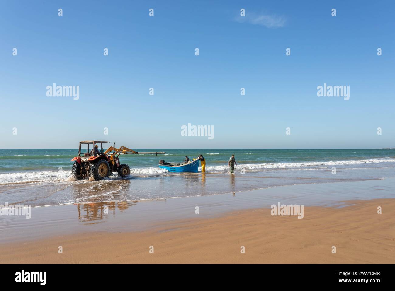 Un tractor está en la playa, listo para levantar un pequeño barco de pesca azul de madera del agua después de un viaje de pesca. Taghazout, Marruecos, Norte de África. Foto de stock