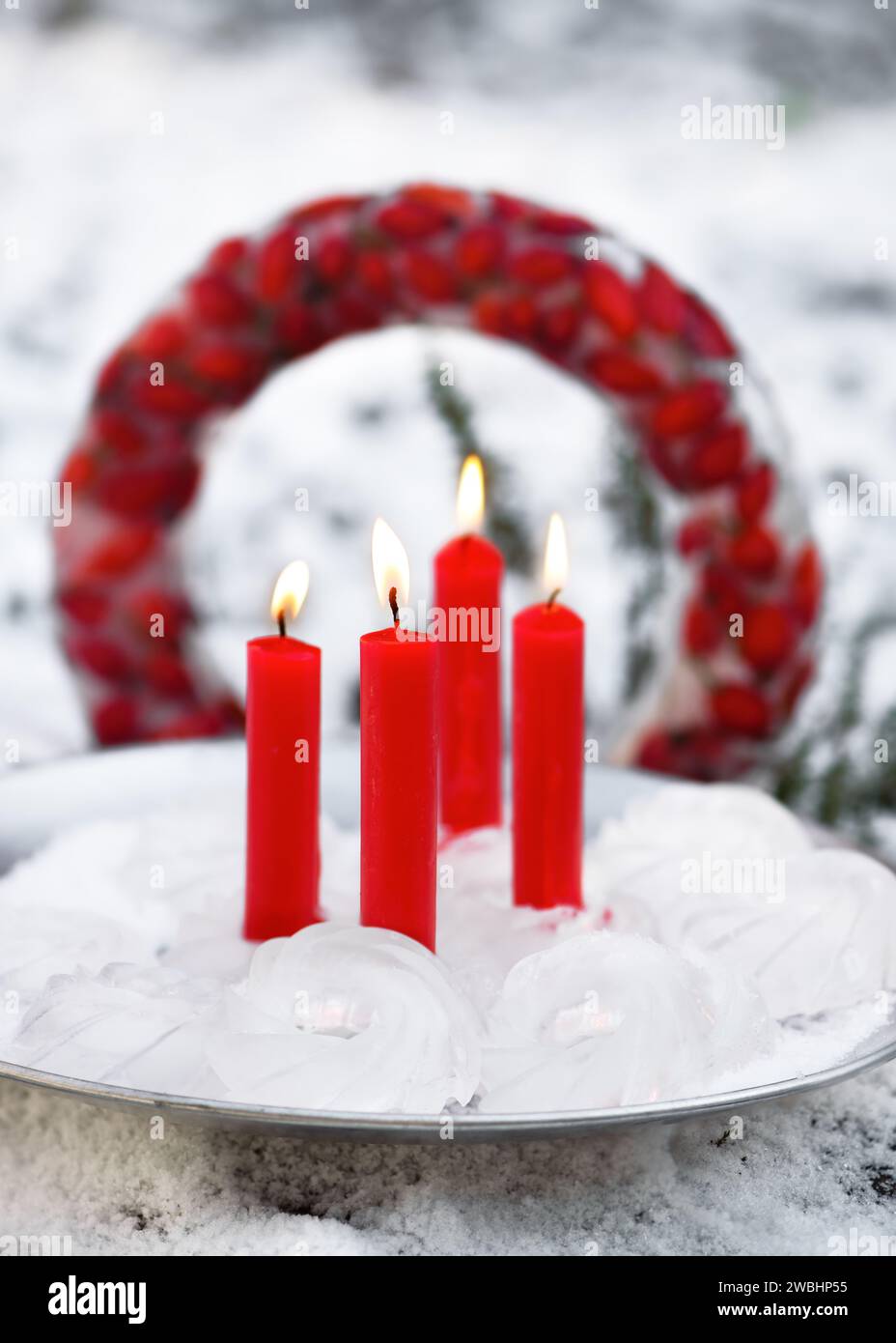 Decoración de hielo de jardín hecha a mano en forma de mini Bundts, con cuatro velas rojas ardientes en placa de metal rústico delante de la corona de Navidad congelada. Foto de stock