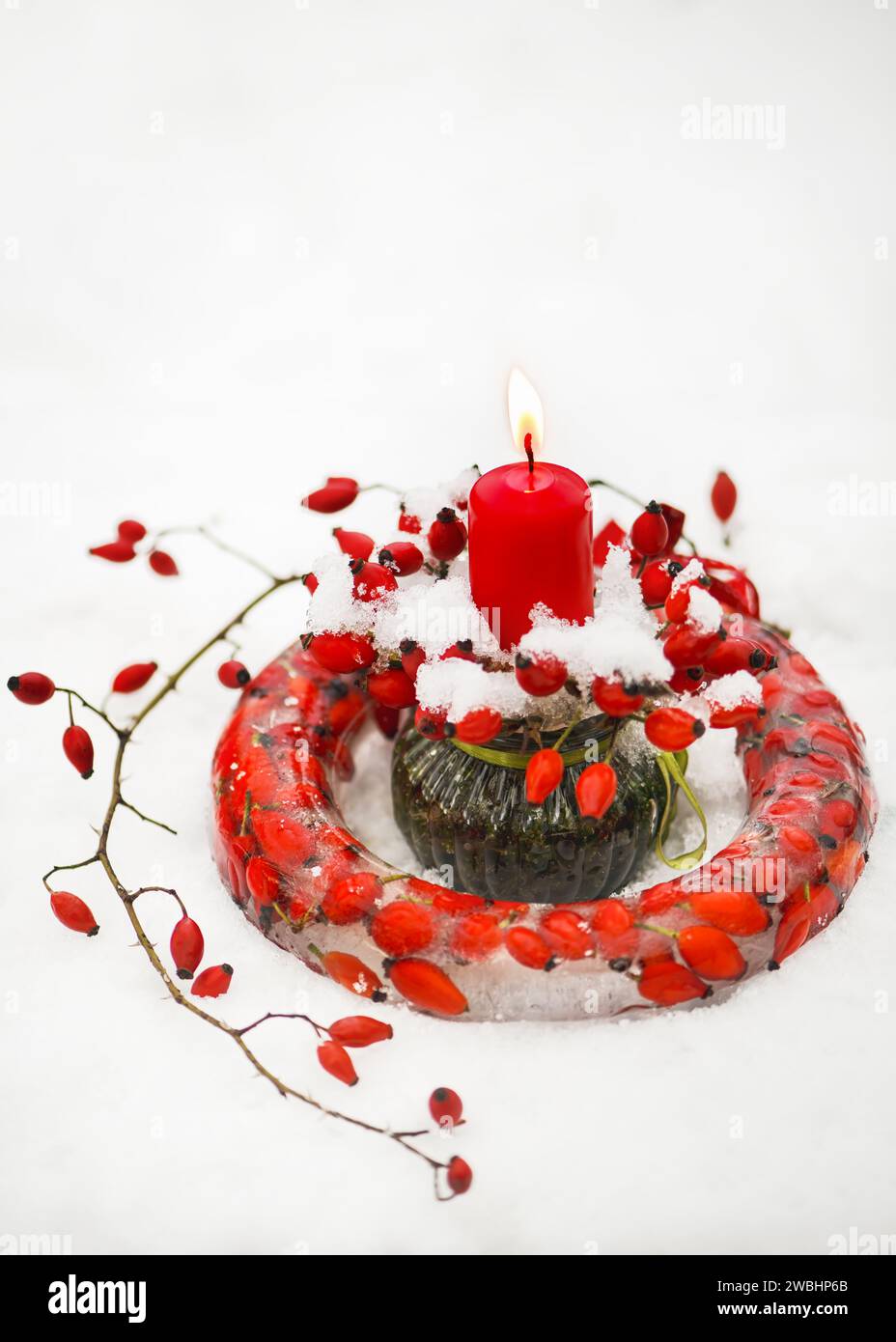 Corona de Navidad congelada hecha a mano en forma de torta de anillo hecha de hielo, moras de caderas de rosa rojas con vela roja ardiente en la nieve. Jardín decoración al aire libre Foto de stock