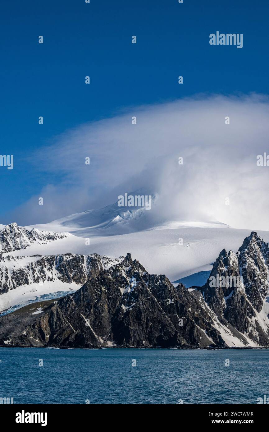 Costa rocosa de la Isla Elefante y altos picos nevados, parcialmente oscurecidos por lenticulares y otras nubes, Monte Pendragón, pico nevado, Foto de stock