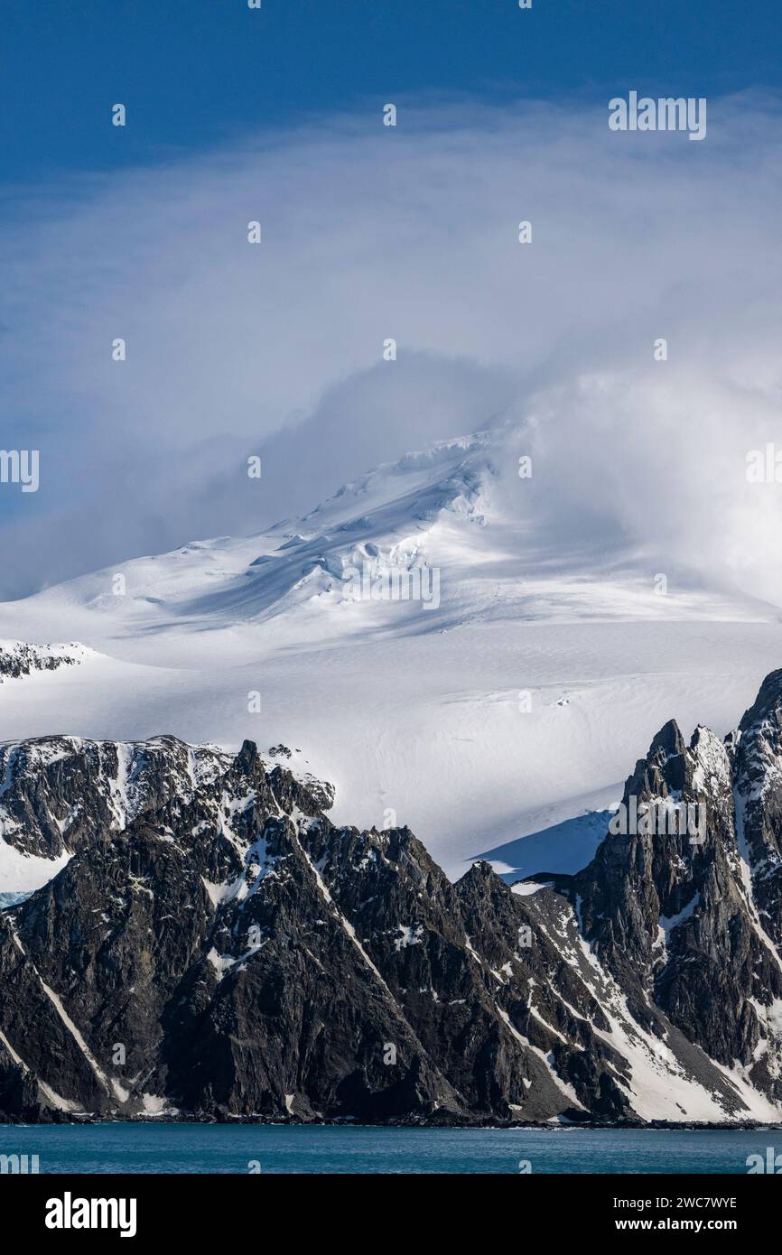 Costa rocosa de la Isla Elefante y altos picos nevados, parcialmente oscurecidos por lenticulares y otras nubes, Monte Pendragón, pico nevado, Foto de stock