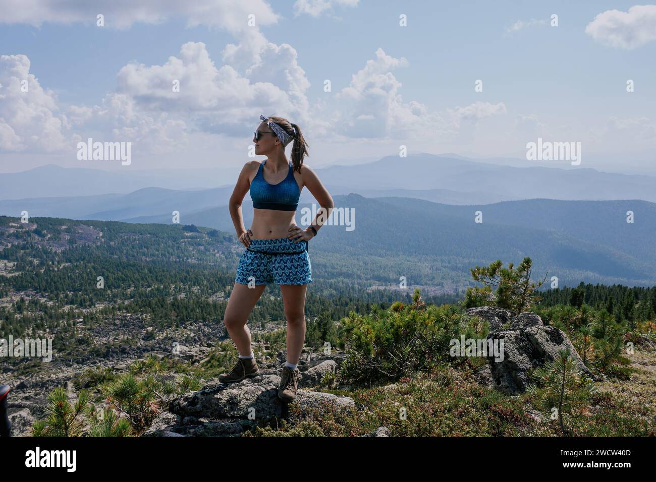 Mujer turista en gafas de sol, pantalones cortos azules y sujetador deportivo se encuentra en una piedra contra el telón de fondo de un paisaje de verano con colinas verdes con coníferas Foto de stock