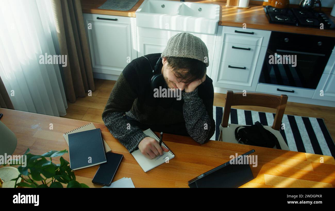 Joven estudiante feliz con gorro sentado en la cocina moderna en casa trabaja con su tableta y toma notas en cuaderno. Tiro de ángulo alto Foto de stock