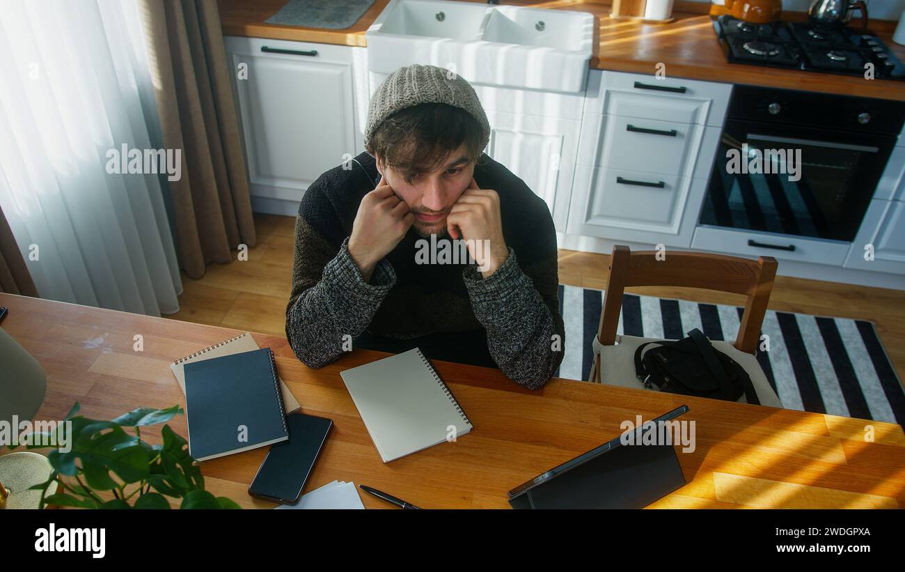 Hombre considerado con gorro sentado en la cocina moderna en casa trabaja con su tableta y toma notas en cuaderno. Tiro de ángulo alto Foto de stock
