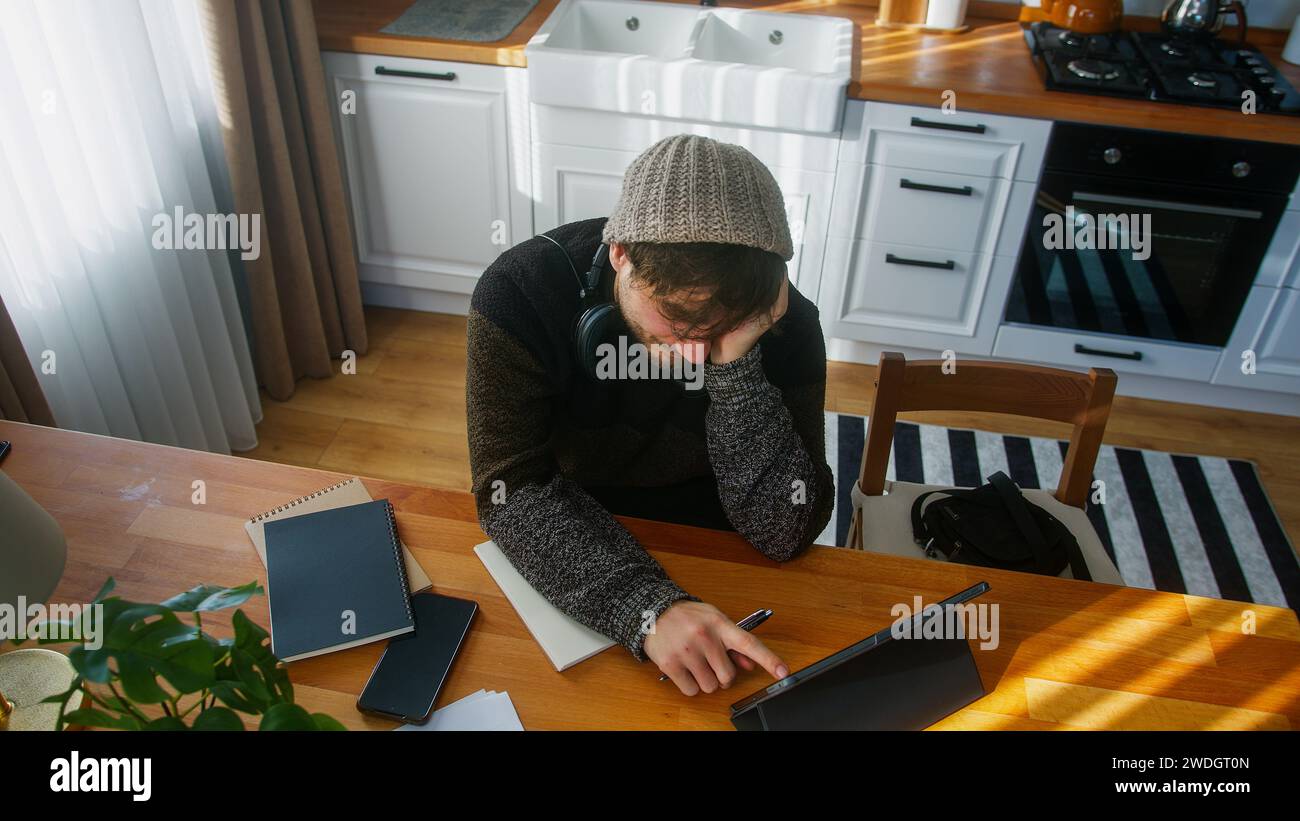 Un estudiante masculino con gorro sentado en la cocina moderna en casa trabaja con su tableta y toma notas en cuaderno. Tiro de ángulo alto Foto de stock