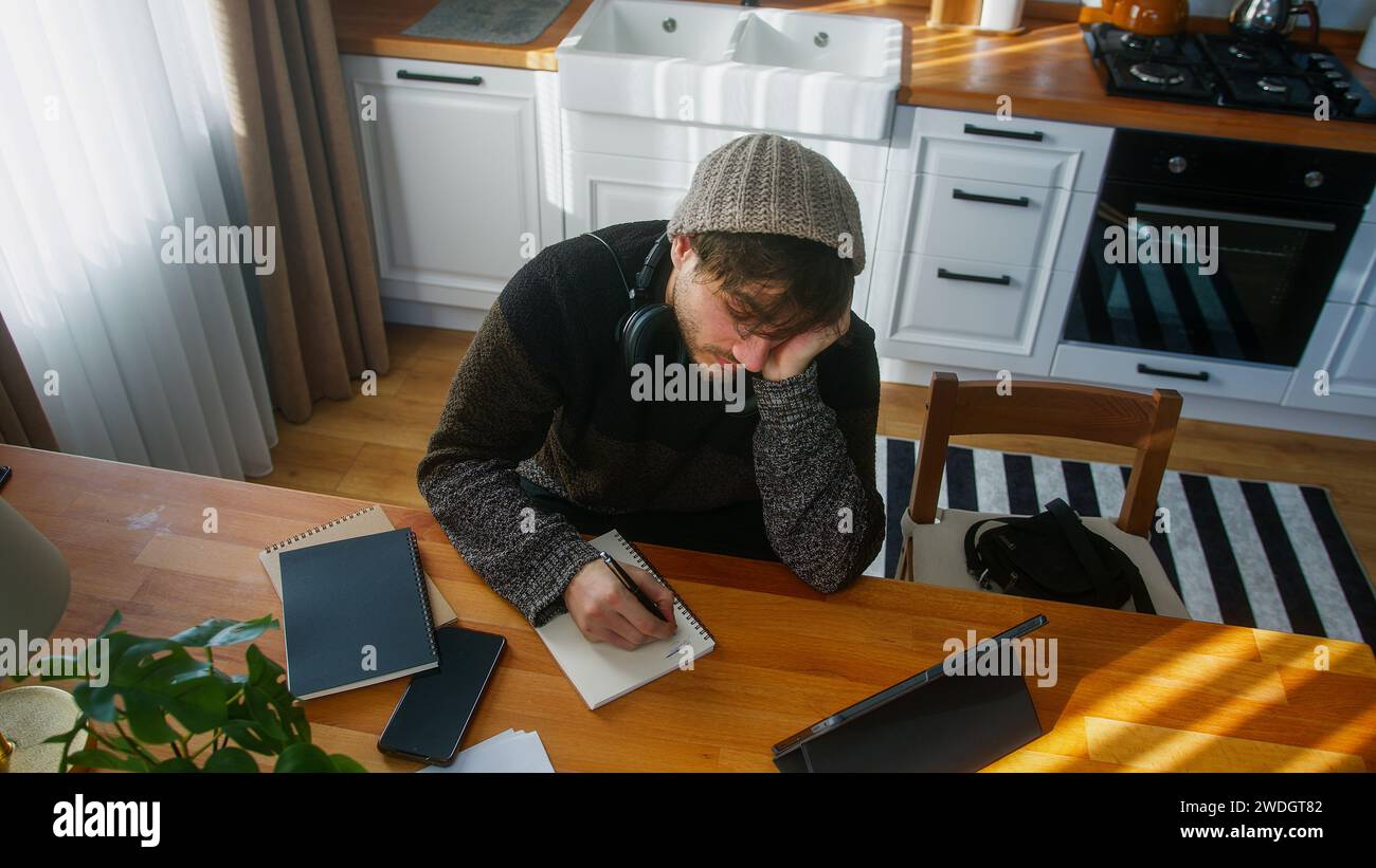 Hombre estudiante aburrido con gorro sentado en la cocina moderna en casa trabaja con su tableta y toma notas en cuaderno. Tiro de ángulo alto Foto de stock
