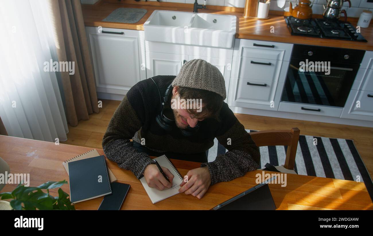 Foto de ángulo alto del joven estudiante con gorro sentado en la cocina moderna en casa trabaja con su tableta y toma notas en cuaderno Foto de stock