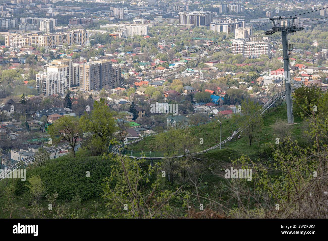 Telesilla vacía que conduce al parque de atracciones Kok Tobe y rodelbahn en la ladera de la montaña. Hierba verde y arbustos, soleado día de primavera. Ver edificios Almaty ciudad Foto de stock