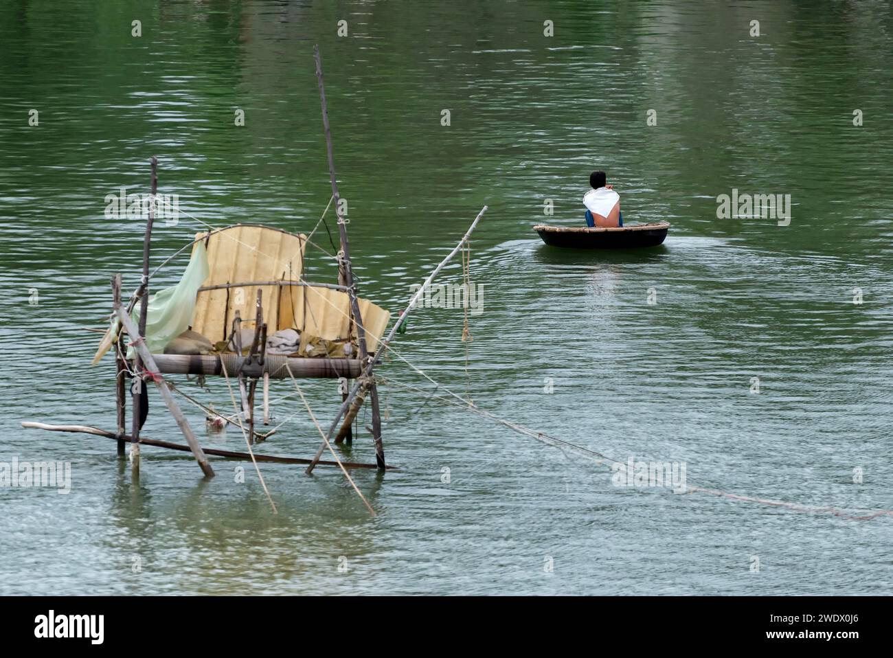 Pescador en bote de cesta o thung chai - barco redondeado hecho de bambú, con cabaña de pesca borrosa a la izquierda en Vietnam Foto de stock
