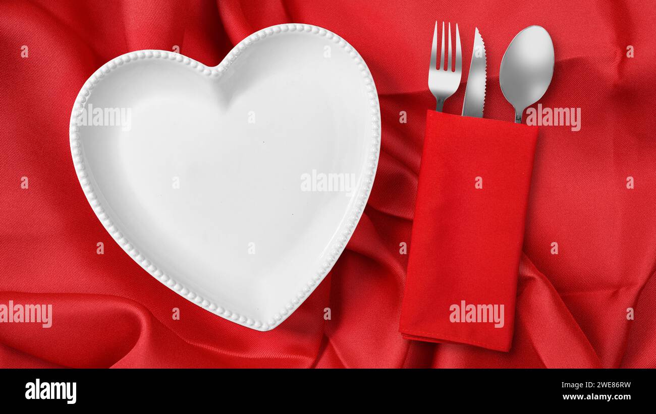 Placa de corazón con cubiertos de tenedor, cuchillo y cuchara en servilleta roja sobre un fondo de servilleta roja. Cena romántica Concepto del Día de San Valentín Foto de stock