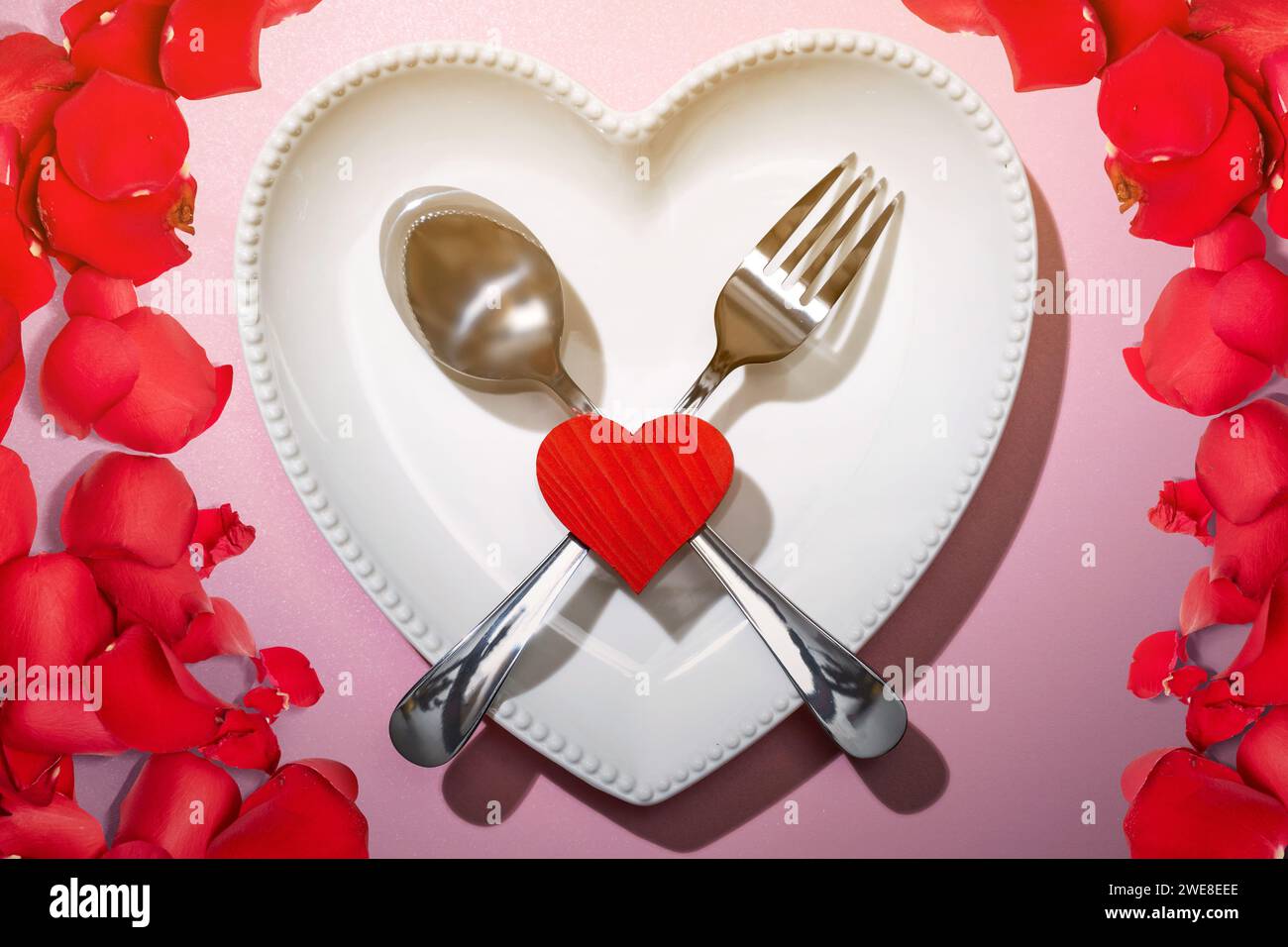 Placa de corazón con cubiertos de cuchara y tenedor con un corazón rojo y pétalo de rosa sobre un fondo rosa. Cena romántica Concepto del Día de San Valentín Foto de stock