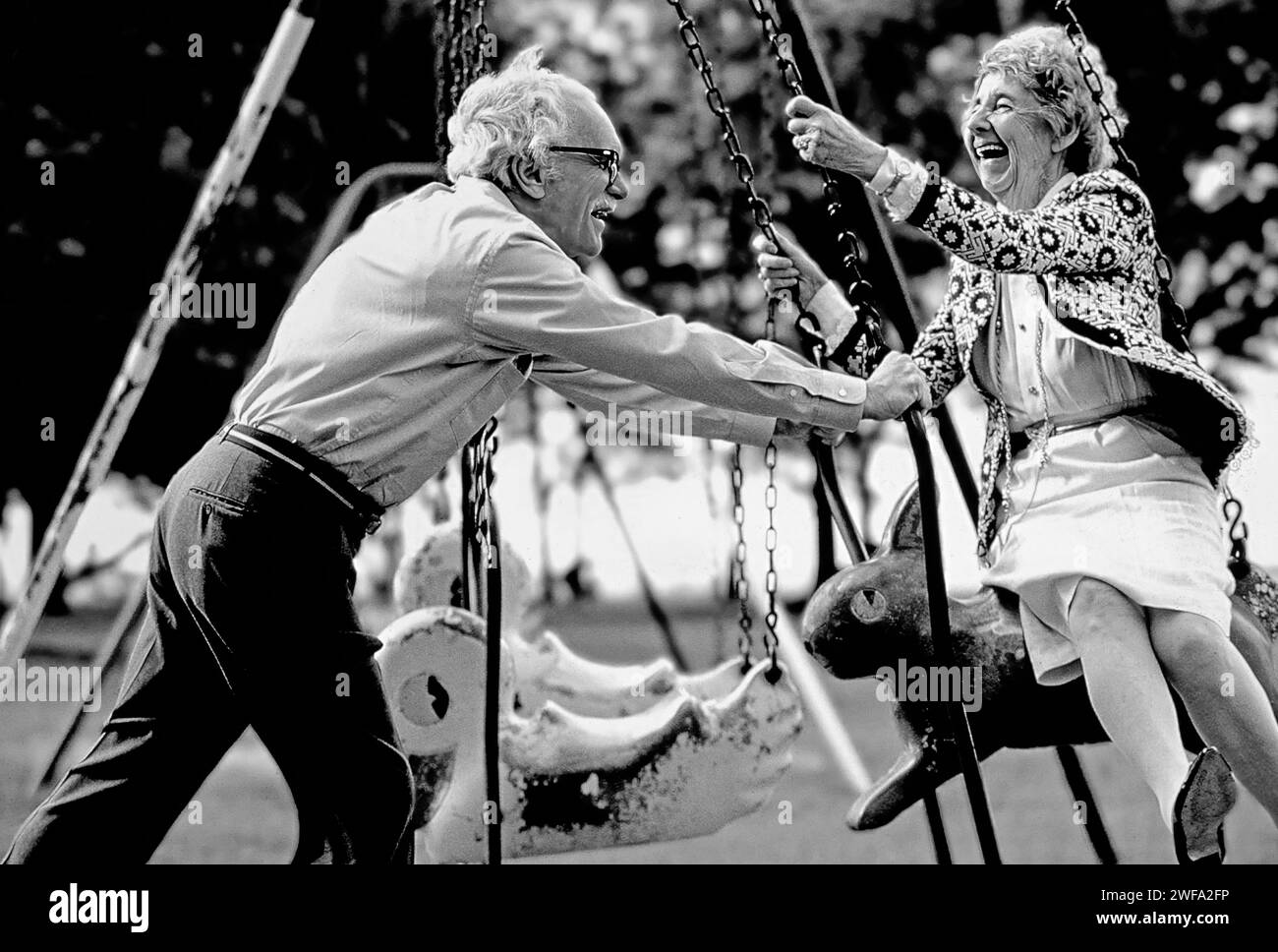 Un hombre y una mujer mayores activos riendo, sonriendo, y disfrutando de un columpio conjunto en el parque Foto de stock
