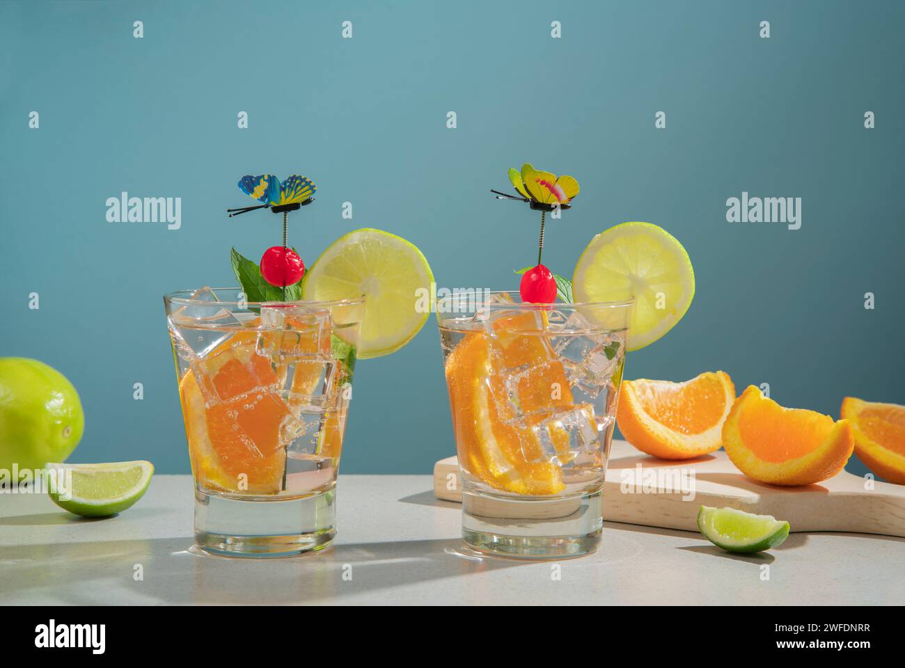 Dos cócteles en vaso de vidrio rellenos de hielo y rebanada de naranja adornada con rodaja de limón, cereza y alfiler de mariposa sobre una mesa blanca con trozos de ora Foto de stock