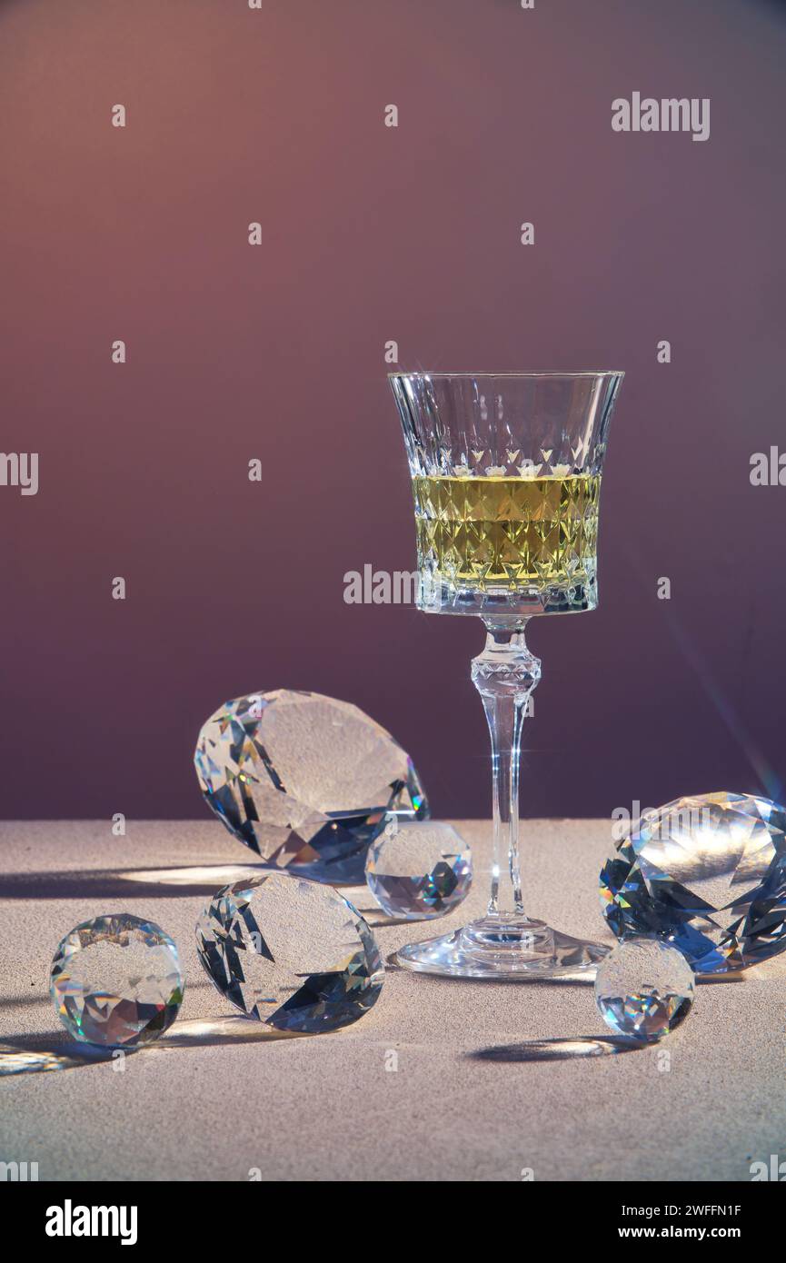Un vaso transparente lleno de vino o bebida amarilla, decorado con diamantes en muchos tamaños en fondo degradado. Concepto de lujo Foto de stock