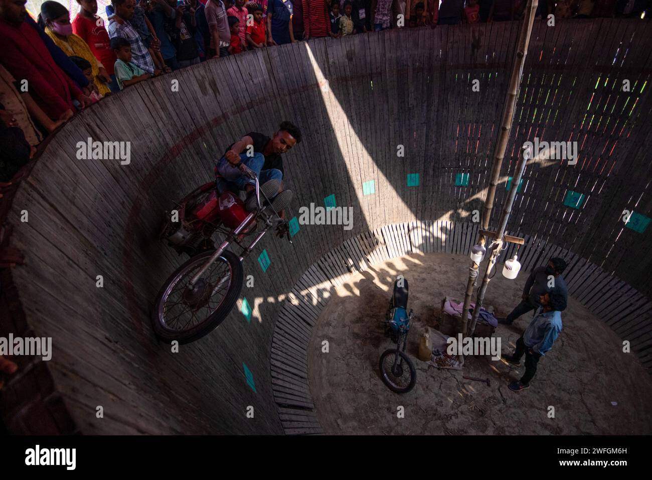 Dhaka, Khulna, Bangladesh. 31 de enero de 2024. Los conductores atrevidos entretienen a los espectadores conduciendo peligrosamente motocicletas en un "muro de la muerte" vertical en Khulna, Bangladesh. Los artistas viajan sin engranajes protectores a velocidades de hasta 80 km/h para que sus vehículos puedan equilibrarse en la tabla de madera de 25 pies de altura. Los jinetes realizan acrobacias que desafían la muerte para sorprender a los transeúntes en un intento de ganar dinero extra mientras viajan verticalmente por el lado de una pared. Actúan para entretener a aquellos que han pagado una cuota de entrada de 20 peniques por el entretenimiento. Hasta 100 espectadores pueden ver a la vez ofreciendo dinero Foto de stock