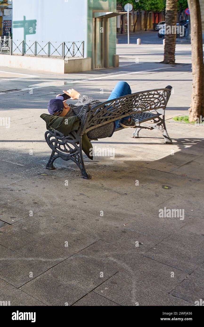 Hombre adulto irreconocible descansando en un banco de parque público y mirando su teléfono celular Foto de stock