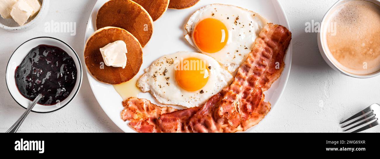 Desayuno con huevos fritos, tocino, panqueques y café, vista superior, banner. Huevos fritos soleados con tocino en el plato y café cappuccino de la mañana. Amer Foto de stock