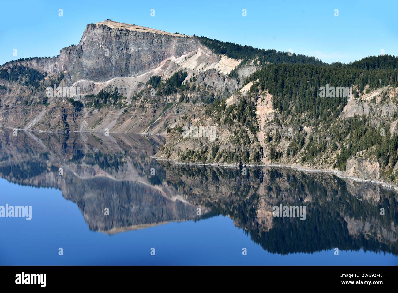 La costa se refleja a la perfección en Crater Lake, Parque Nacional Crater Lake, en Oregón. Acantilados empinados y cielo azul se reflejan. Foto de stock