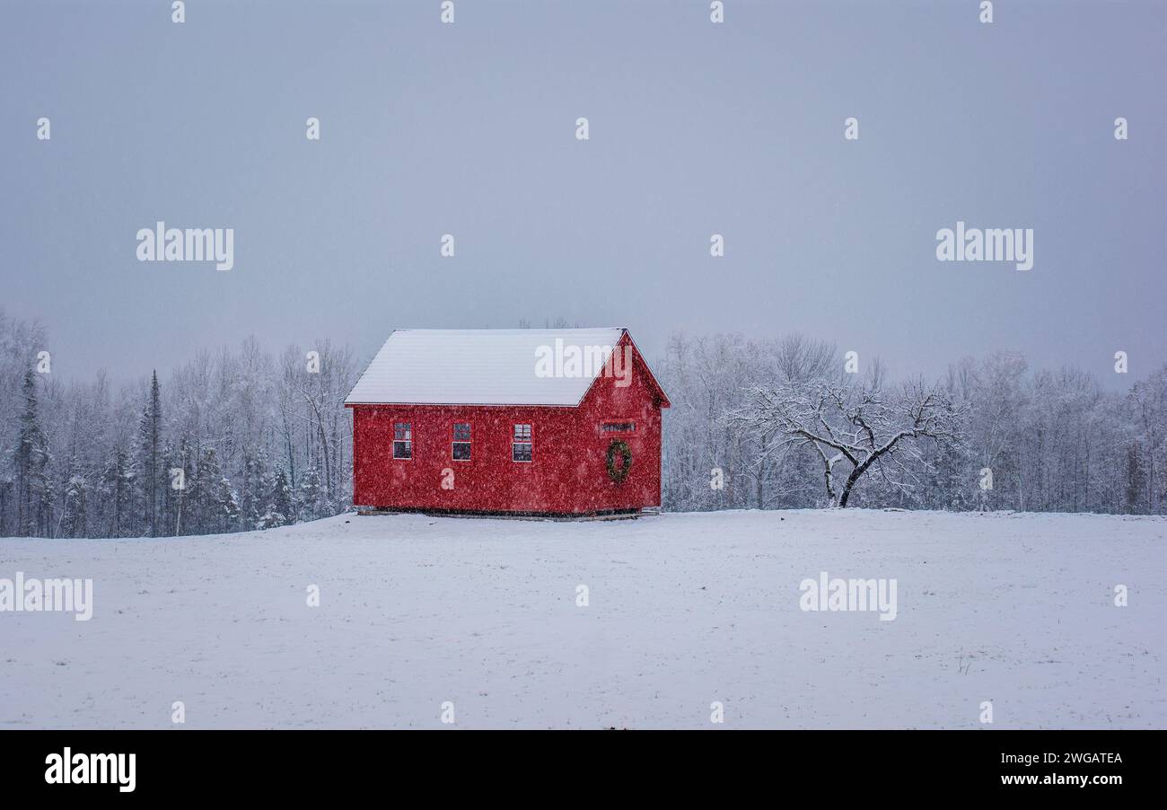 Una encantadora cabaña roja con decoraciones navideñas festivas, bajo fuertes nevadas, en un paisaje invernal. Monson, Maine, Estados Unidos Foto de stock