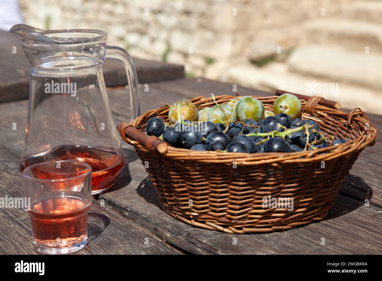 Jarra y copa de vino rosa junto a una cesta llena de uvas negras y ciruelas verdes Foto de stock