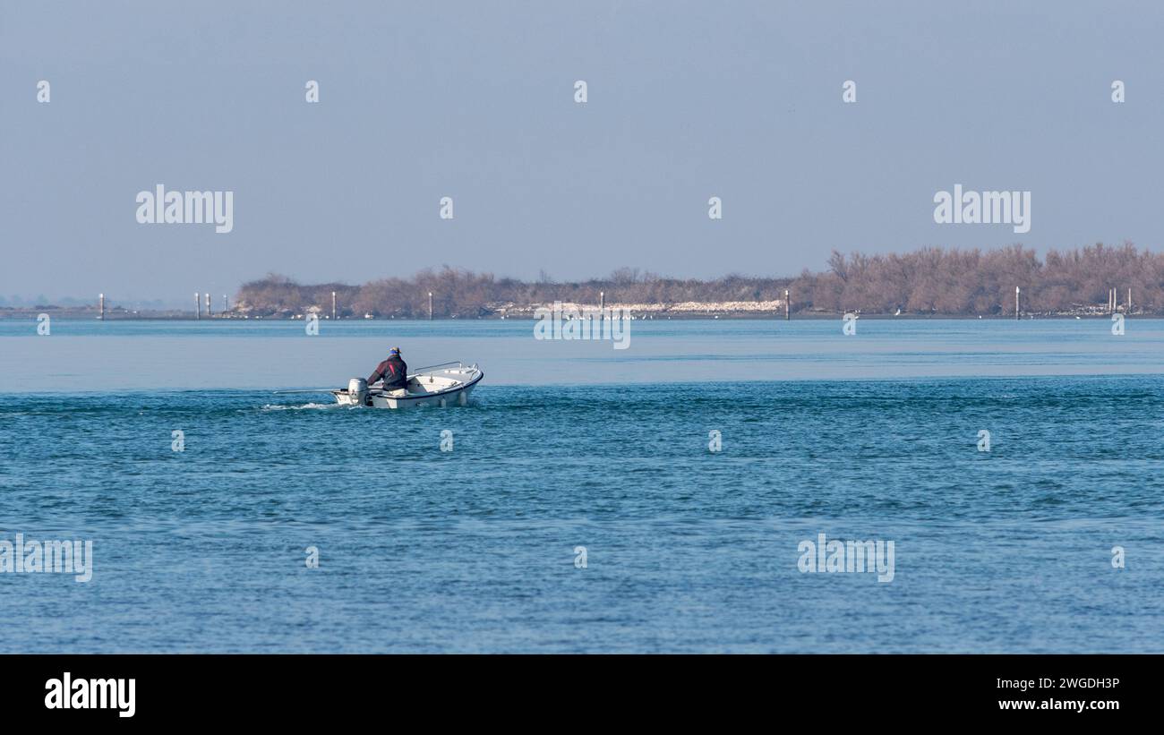 Un pequeño barco pesquero con pescador de edad avanzada a bordo regresa al puerto después de un viaje de inspección en un día de invierno nebuloso en Grado, Italia. Foto de stock