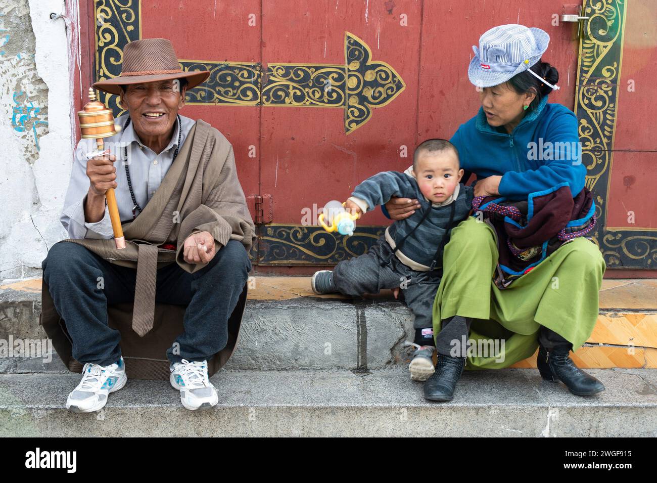 Un hombre que sostiene una rueda de oración tibetana se sienta en escaleras de piedra con su esposa e hijo en la plaza Barkhor, Lhasa. Foto de stock