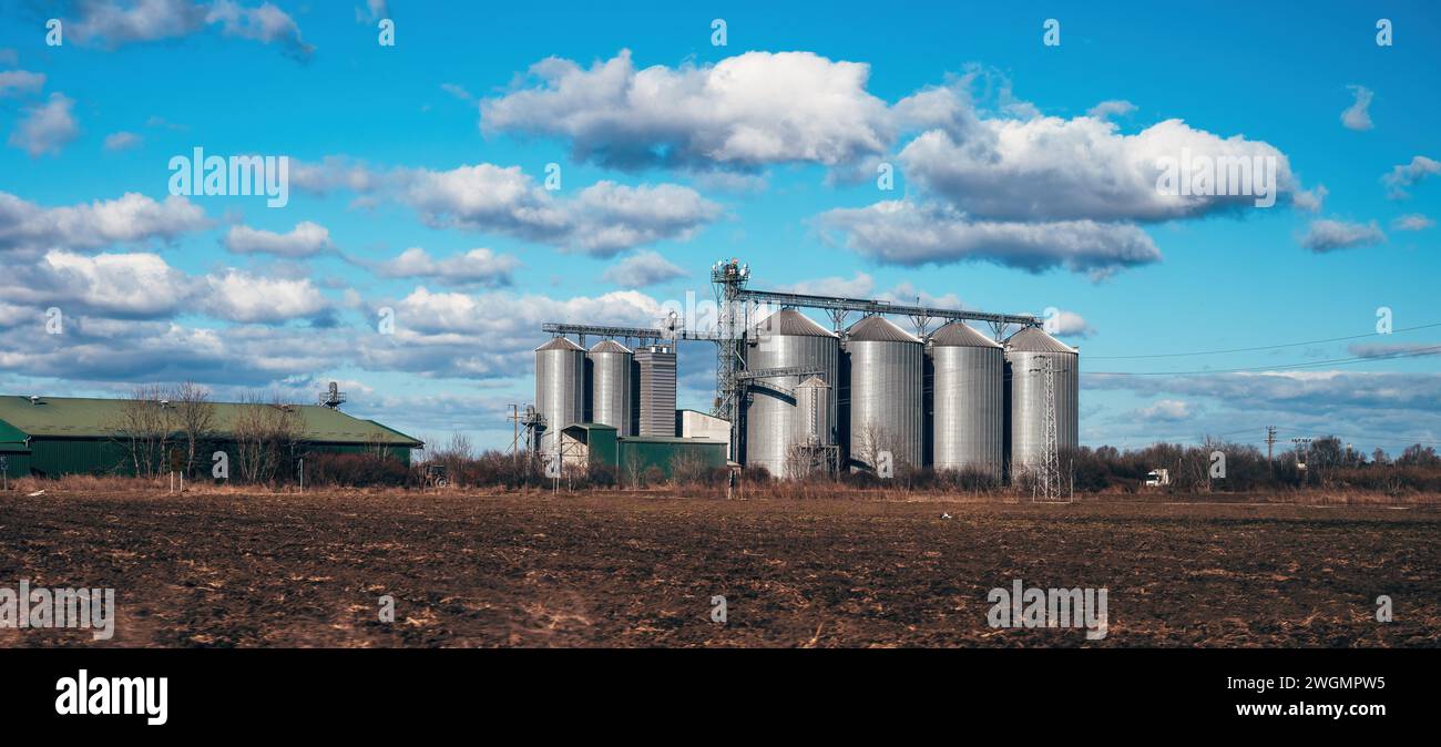 Silos agrícolas de planta de procesamiento de granos rodeados de campos arados, agricultura y edificios agrícolas con hermoso cielo en el fondo Foto de stock