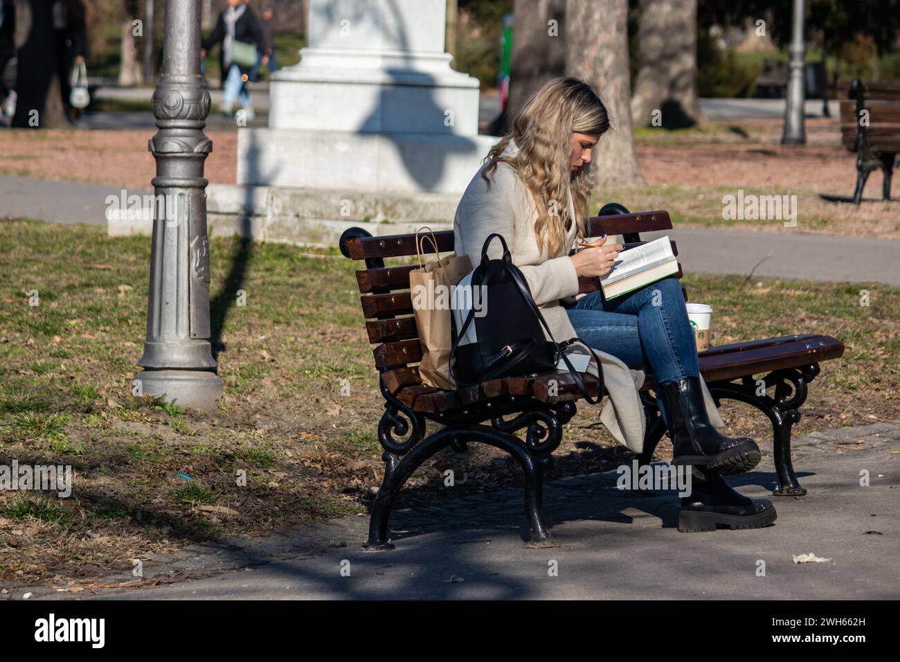 Mujer joven sentada en el banco del parque público en día soleado, disfrutando del buen tiempo y leyendo un libro interesante Foto de stock