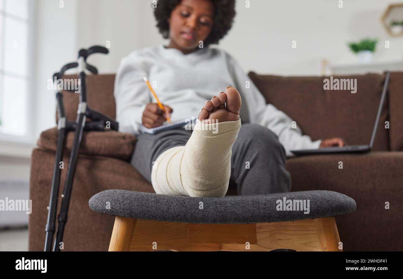 Hermosa, dulce mujer afroamericana trabaja en casa en la sala de estar con computadora portátil y hace notas en el cuaderno. Trabajo remoto con pierna vendada rota Foto de stock