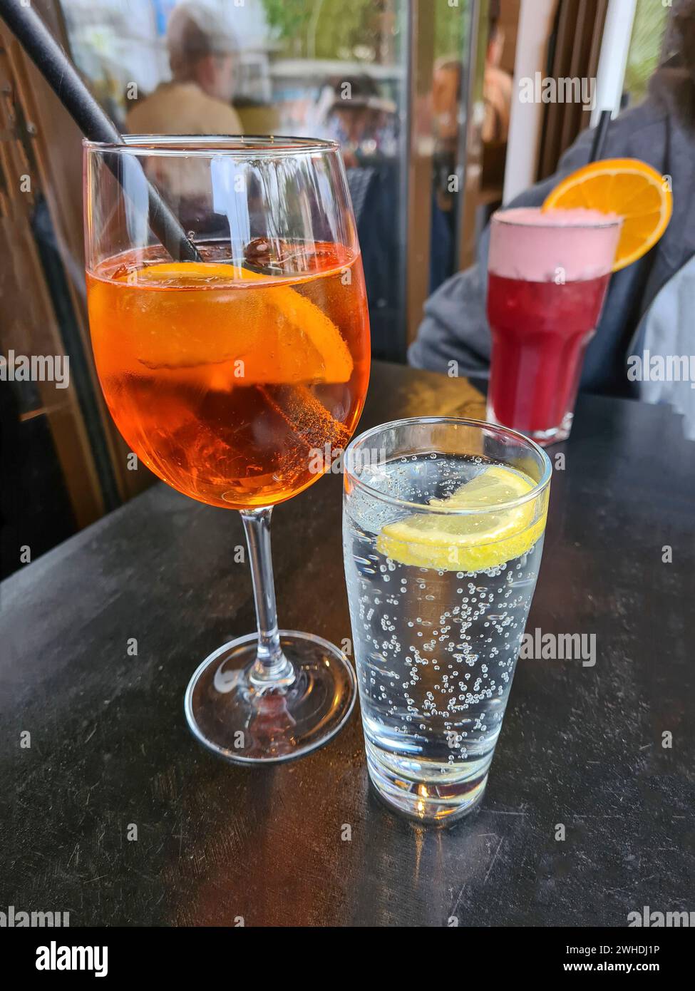 El refresco alcohólico Aperol Spritz se coloca en una mesa junto a un vaso de agua mineral con gas y un cóctel de frutas rojas sin alcohol en el fondo Foto de stock