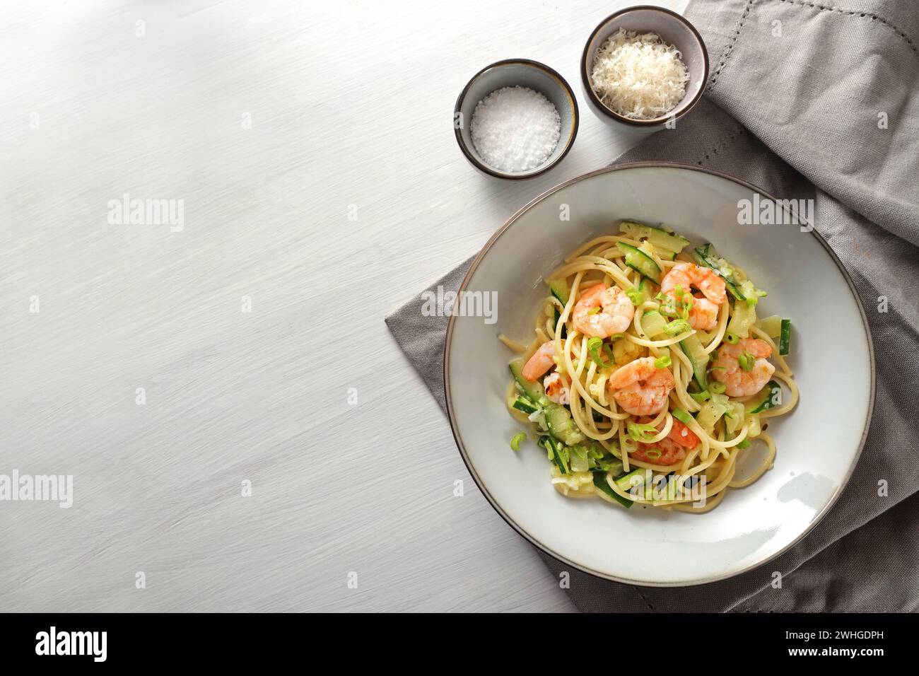 Espaguetis con calabacines calabacines, cebolletas y gambas en un plato gris sobre una mesa pintada de blanco, servilleta y pequeña sal y. Foto de stock