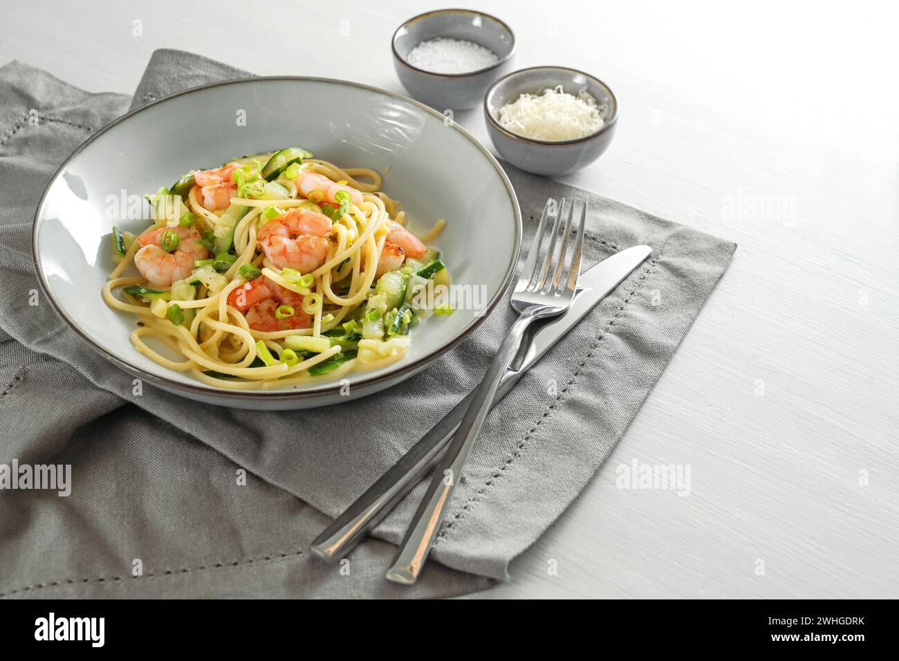 Espaguetis con calabacines calabacines, cebolletas y gambas en un plato gris sobre una mesa blanca con servilleta y cubiertos, saludable Foto de stock