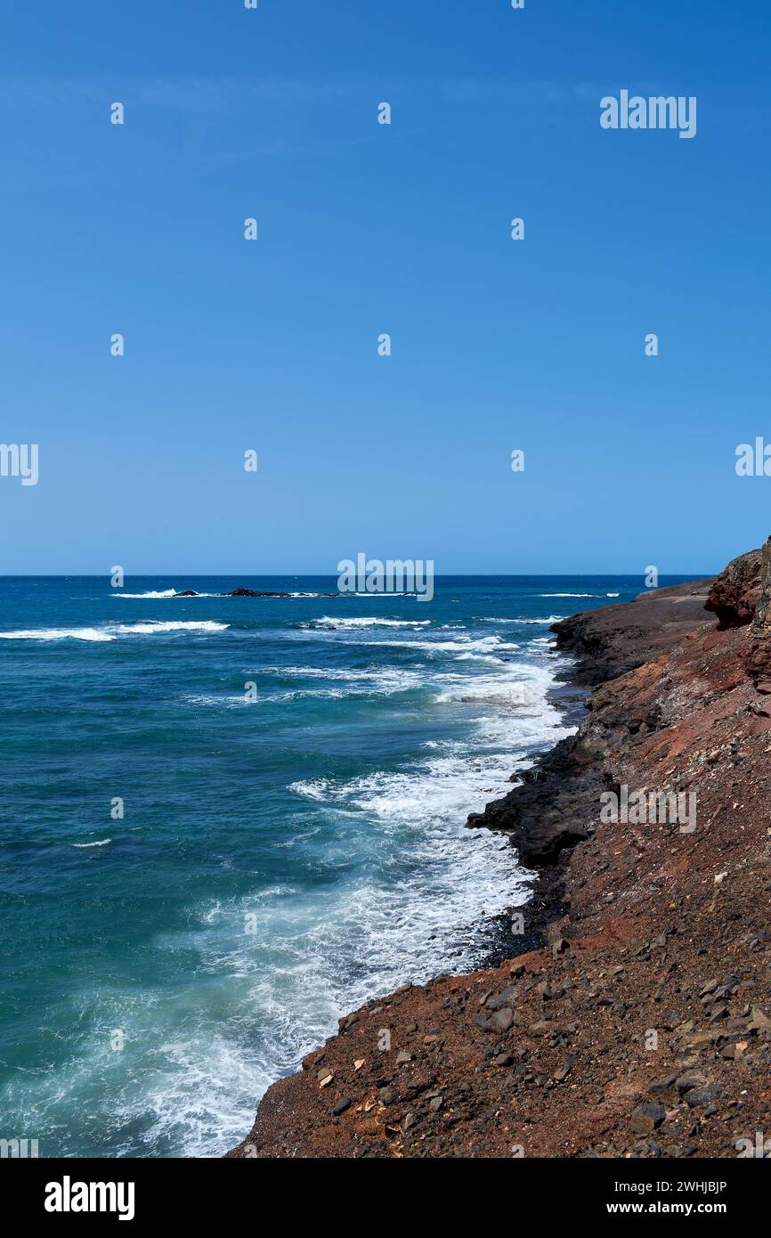 El Océano Atlántico en Jandía con cielo claro y montañas en la parte posterior y olas rompiendo en la orilla Foto de stock