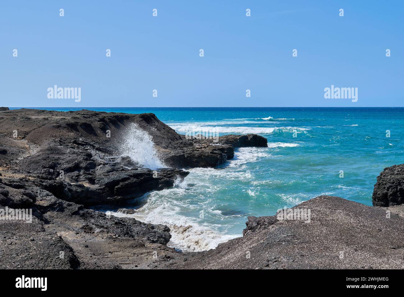 El Océano Atlántico en Jandía con cielo claro y montañas en la parte posterior y olas rompiendo en la orilla Foto de stock