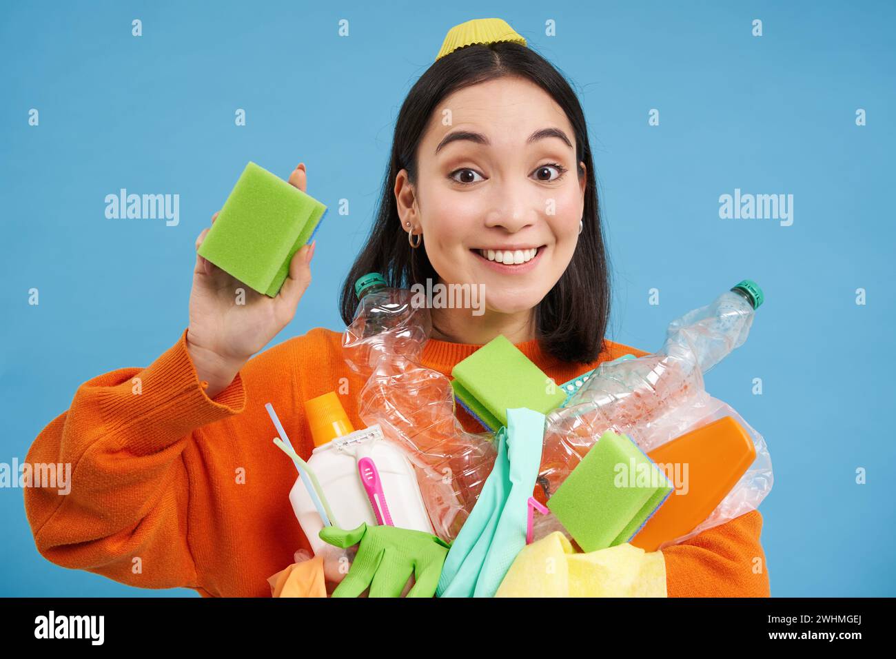 Sonriente entusiasta activista ecológica femenina, sosteniendo esponja de limpieza, botellas de plástico vacías y basura, clasificando los residuos para reciclar Foto de stock