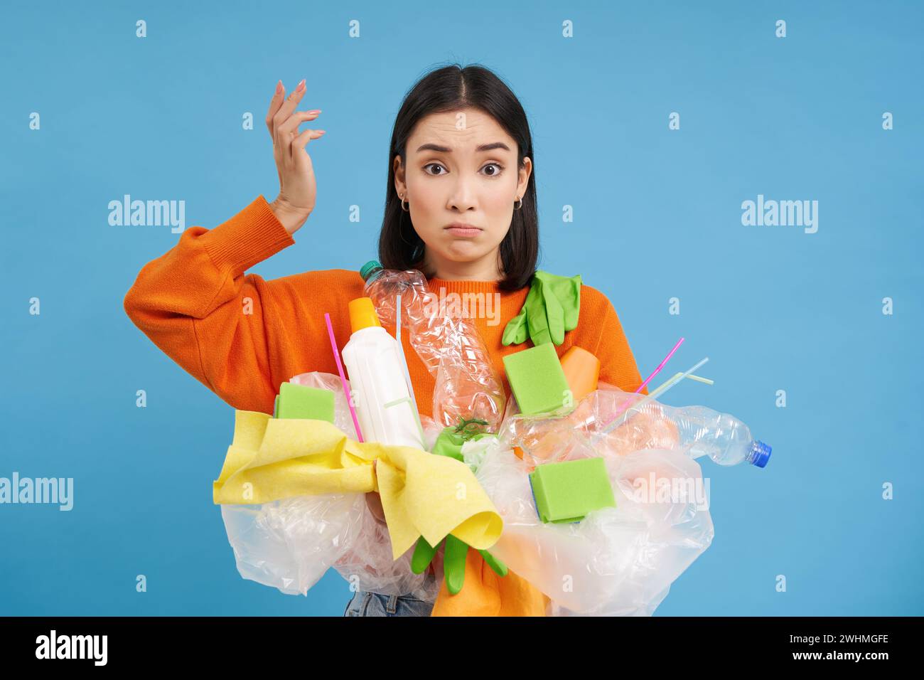 La mujer coreana confundida trata de ponerse verde, reciclar artículos de plástico para el hogar, sostener botellas y guantes, reciclar, backg azul Foto de stock