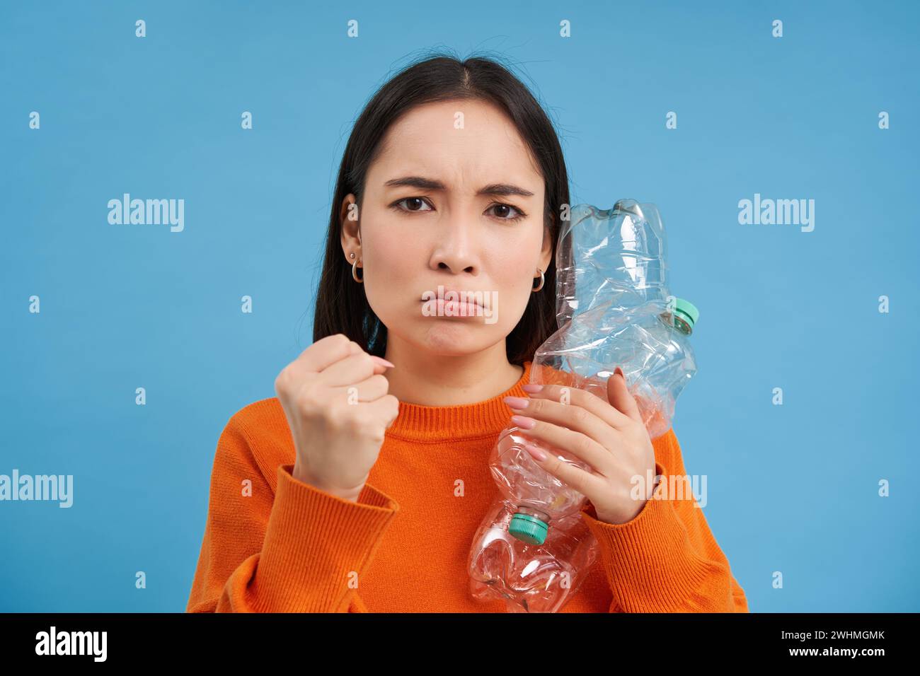 Mujer asiática enojada que sostiene dos botellas de plástico con cara fruncida, chica con estilo de vida verde ecológico, insta a reciclar los desechos, ba azul Foto de stock