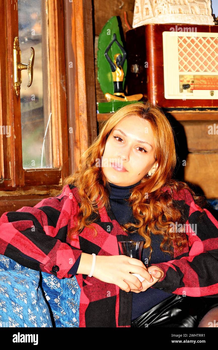 Hermosa joven morena sentada cerca de la ventana y bebiendo té. Ella está sentada en una clásica casa de pueblo turco de madera. mira afuera por ventana Foto de stock
