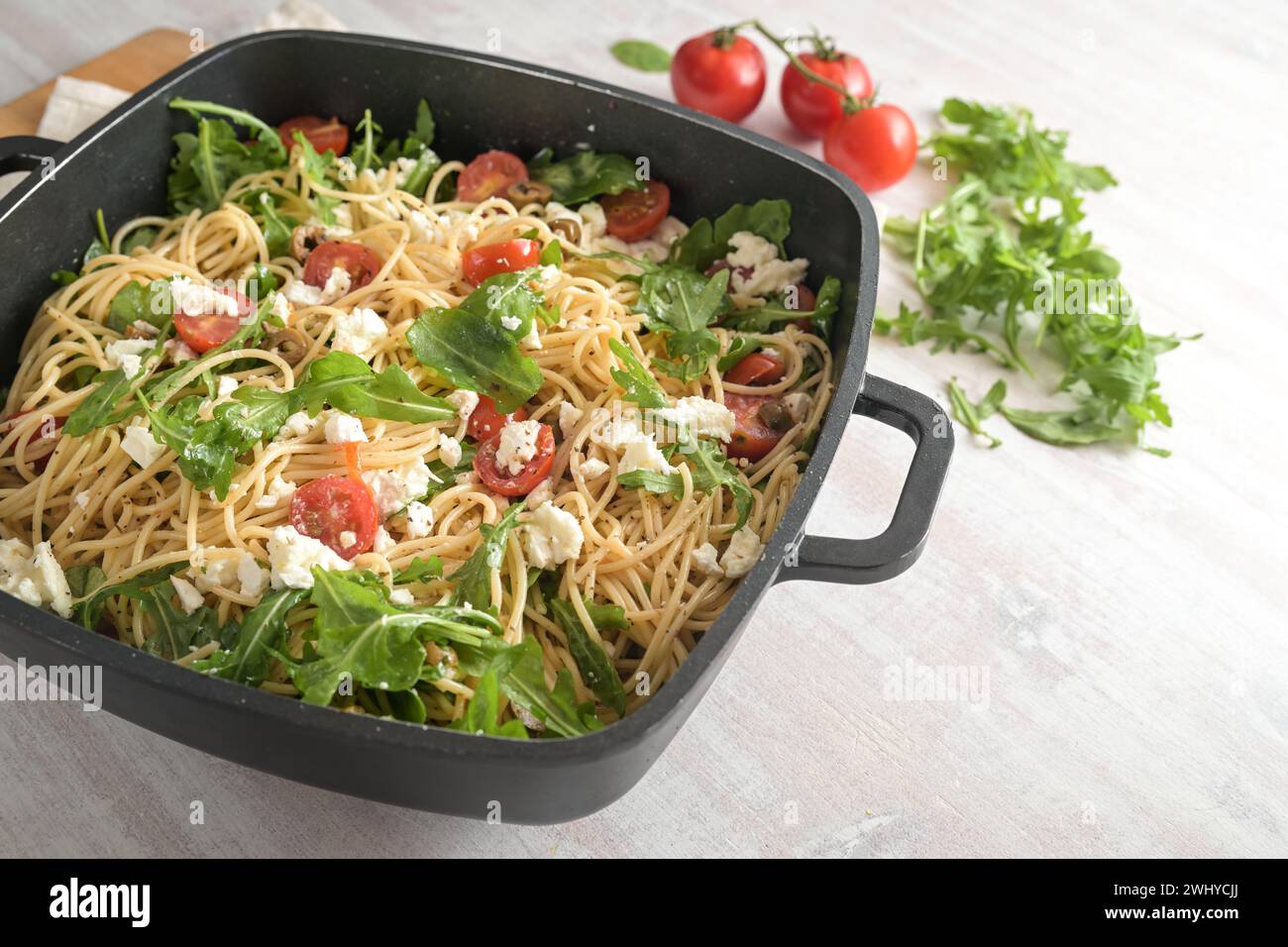 Ensalada de espaguetis con tomates, rúcula, queso mozzarella y aceitunas en un tazón negro grande, comida fría de fiesta mediterránea en un w Foto de stock