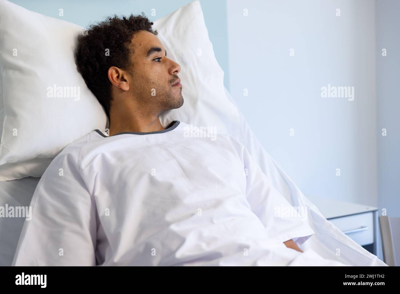 Paciente biracial masculino acostado en la cama en la habitación soleada del hospital Foto de stock