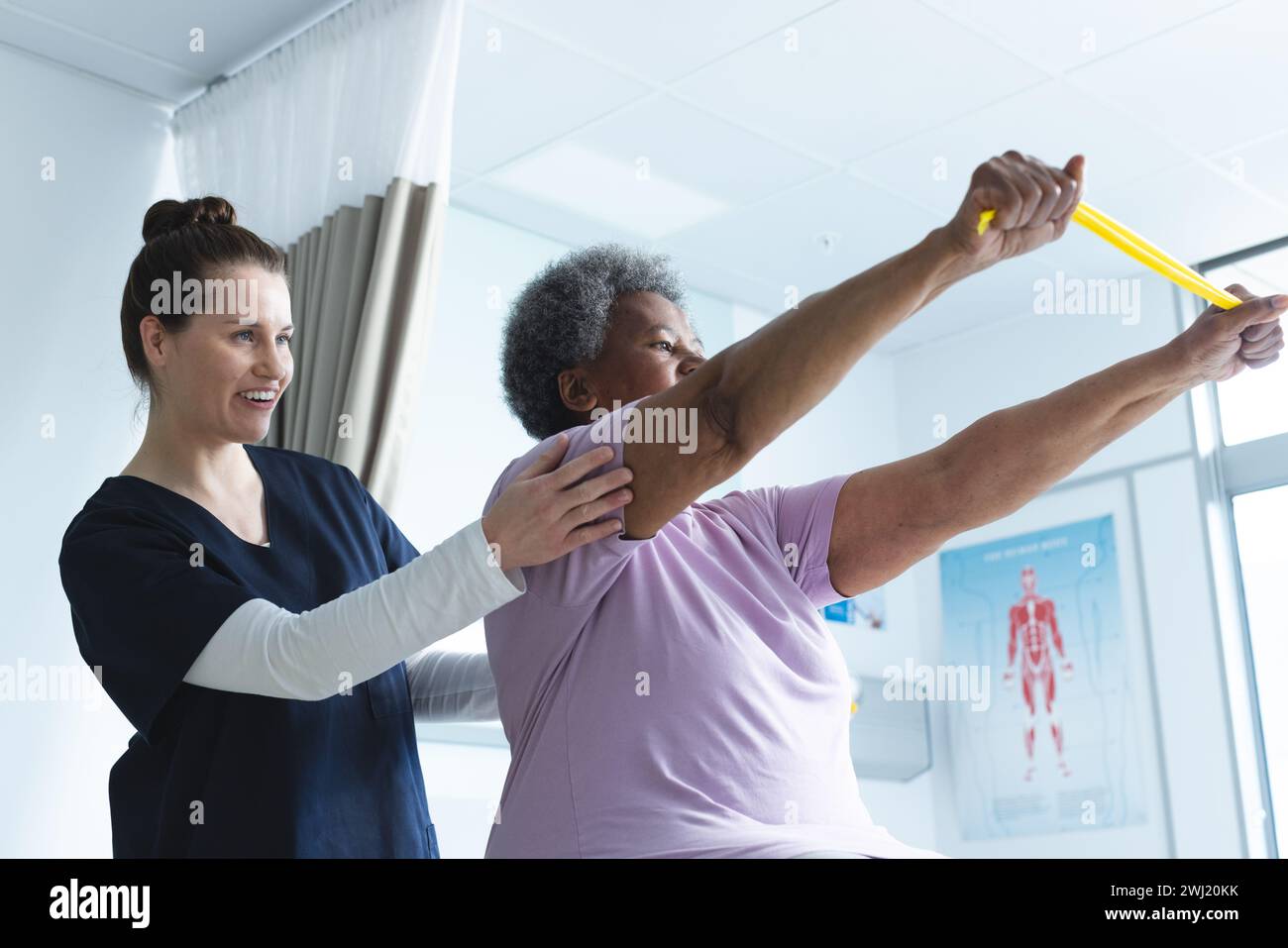 Diversa paciente femenina mayor ejercitando con banda y médico femenino asesorando en habitación de hospital Foto de stock