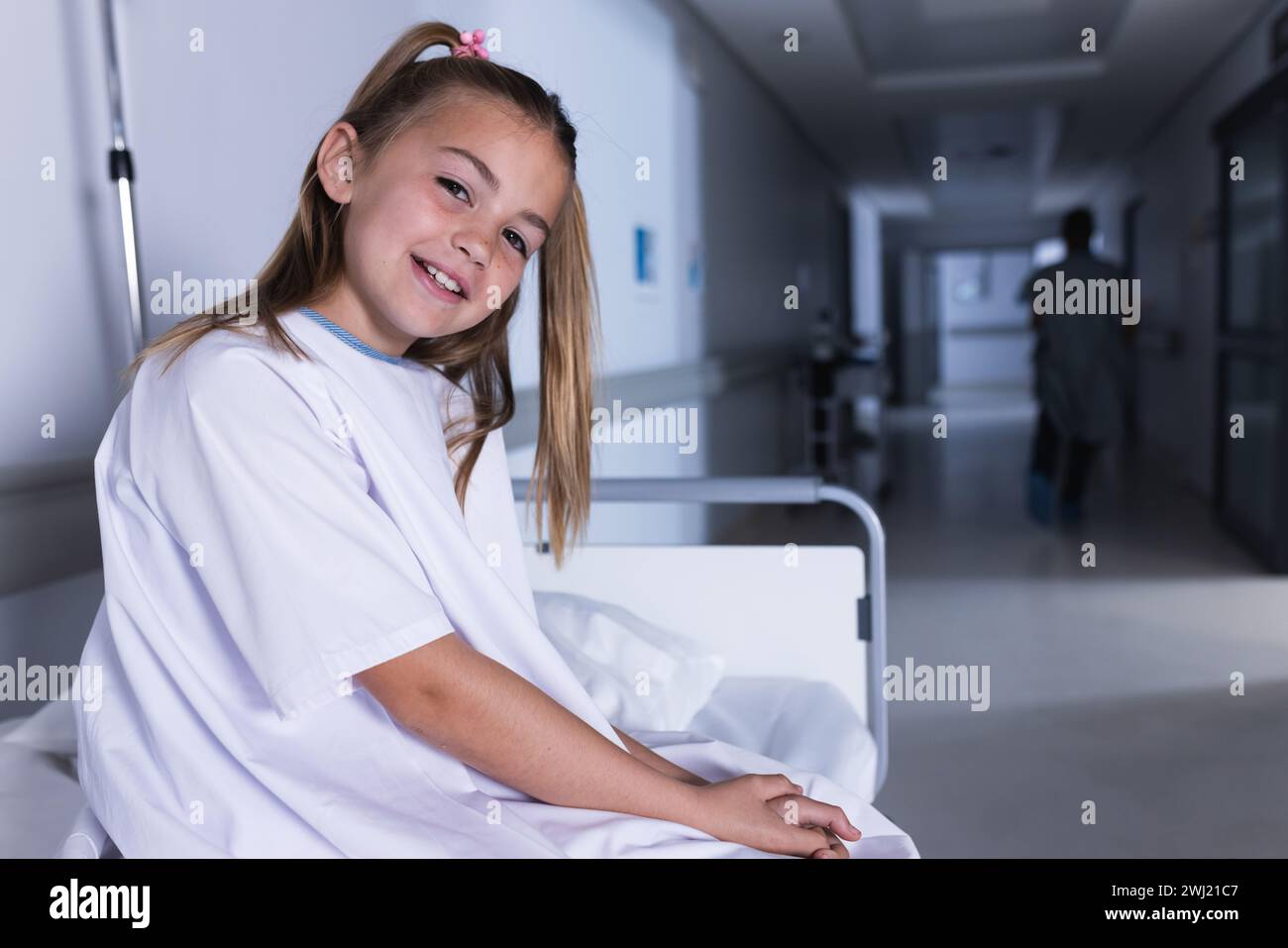 Chica caucásica feliz que se sienta en la cama en el pasillo del hospital con espacio de la copia Foto de stock