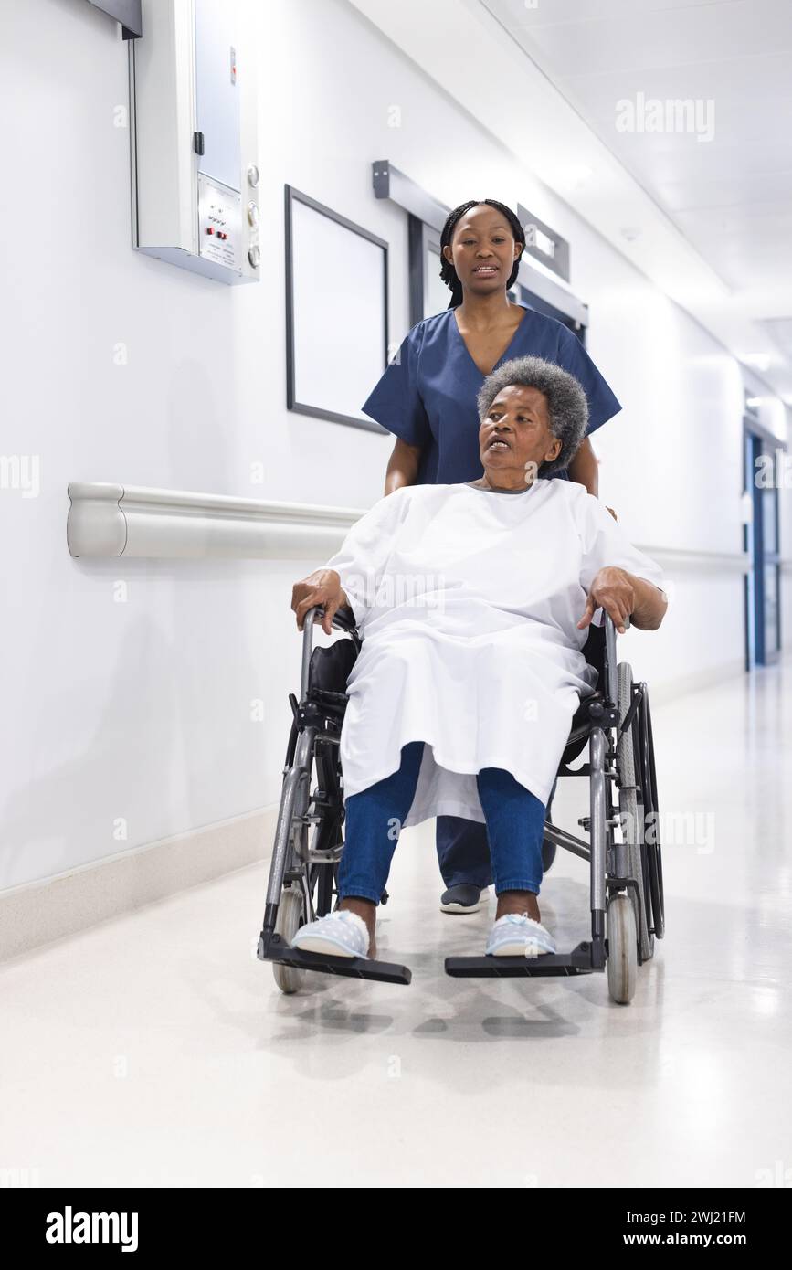 Médico femenino afroamericano caminando con paciente femenino mayor en silla de ruedas en pasillo del hospital Foto de stock