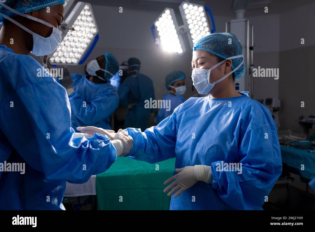 Médico femenino diverso que ayuda a usar guantes protectores en el quirófano del hospital Foto de stock