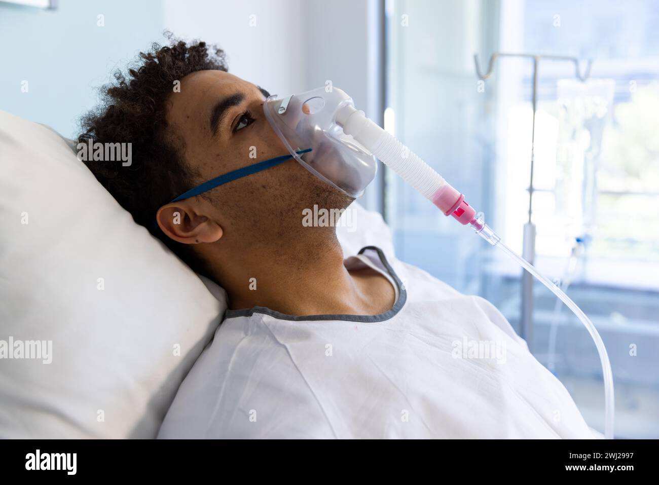 Paciente biracial masculino con máscara de oxígeno acostado en la cama en la habitación soleada del hospital Foto de stock
