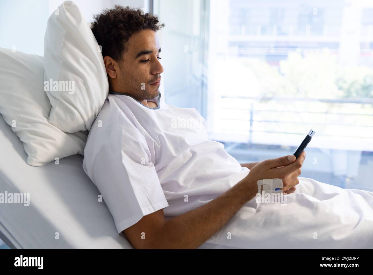 Paciente masculino biracial usando smartphone con goteo iv en cama en habitación de hospital soleado Foto de stock