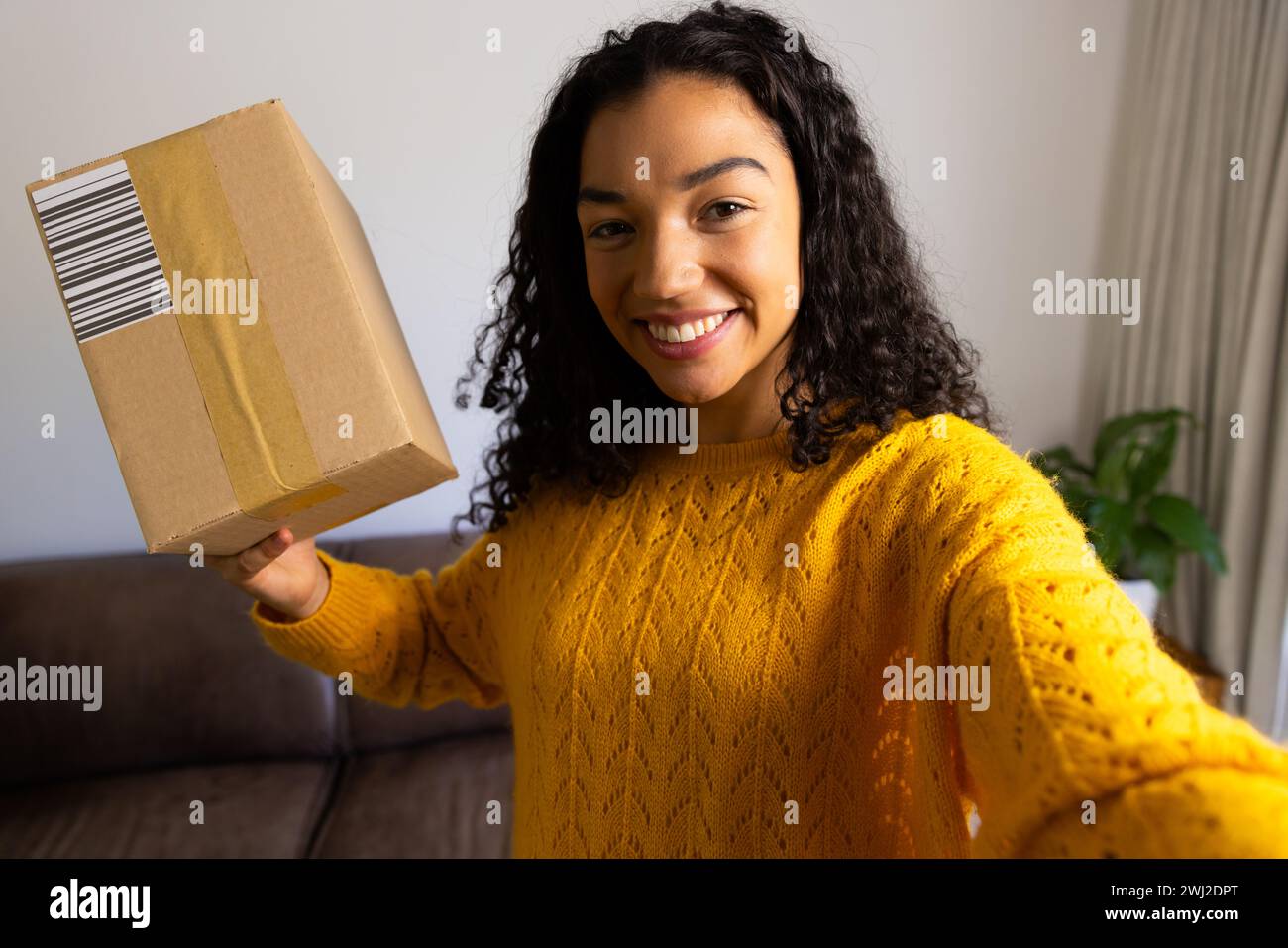 Mujer biracial feliz en suéter amarillo que sostiene el paquete en casa Foto de stock