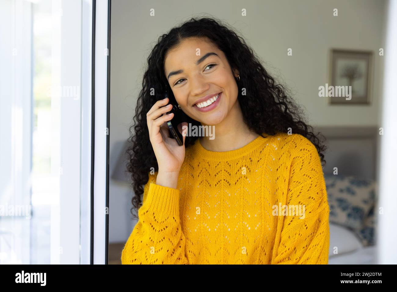 Mujer biracial feliz en suéter amarillo hablando en smartphone en casa Foto de stock