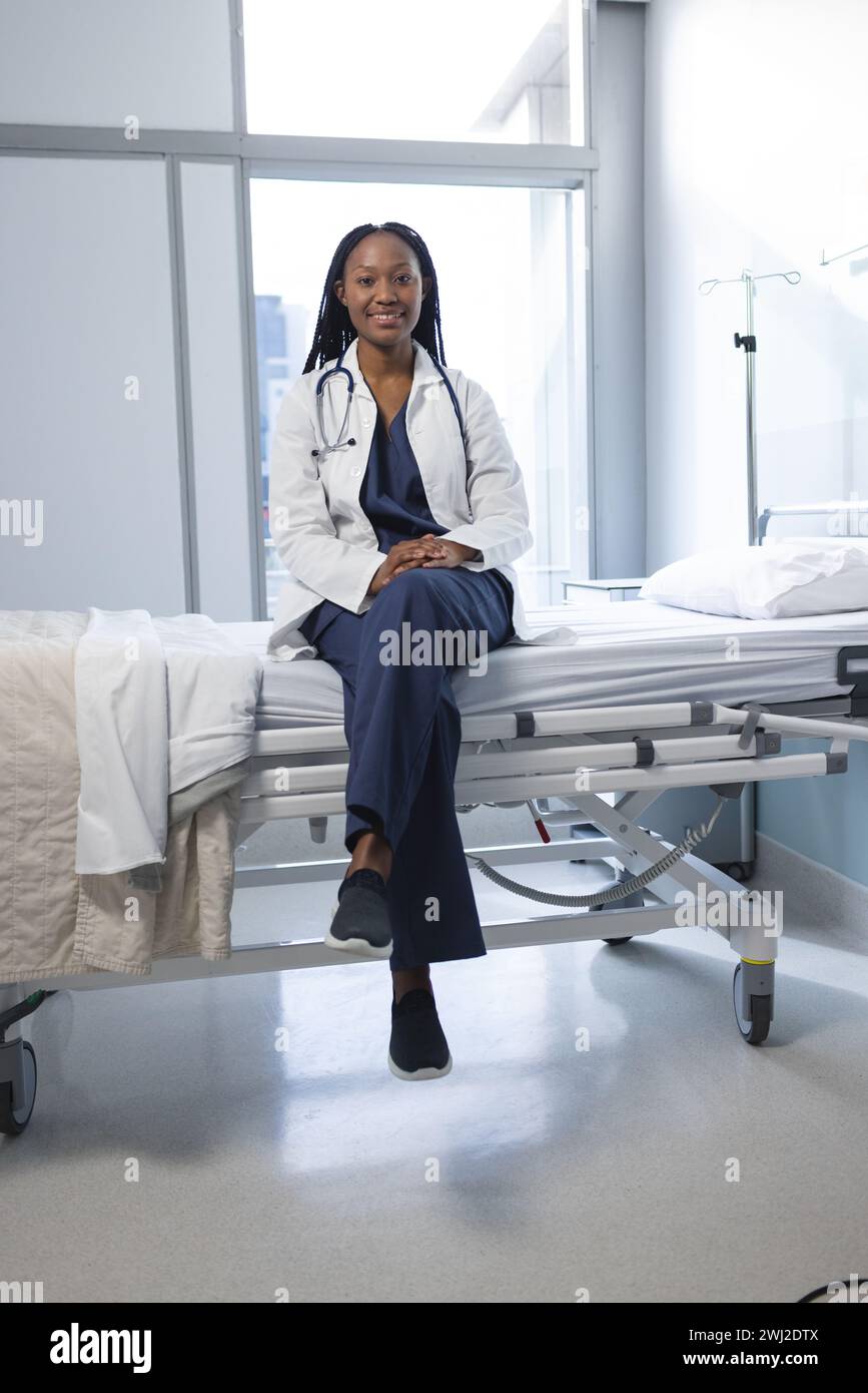 Retrato del médico femenino afroamericano feliz que lleva la capa de laboratorio sentado en la cama en la habitación del hospital Foto de stock