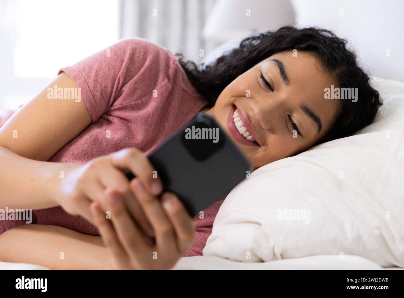 Mujer biracial feliz usando smartphone acostado en la cama en casa soleada Foto de stock